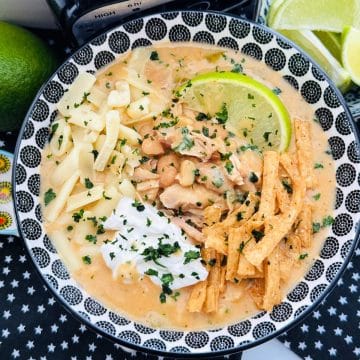 a bowl full of white chicken chili topped with sour cream, a lime wedge, shredded cheese and tortilla chips