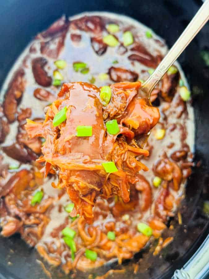 serving a spoonful of bourbon chicken from the crockpot, garnished with green onions